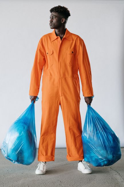 A young man dressed in an orange work jumpsuit stands indoors against a plain light gray background, holding two large blue trash bags filled with waste, with one in each hand. The bags appear to be m