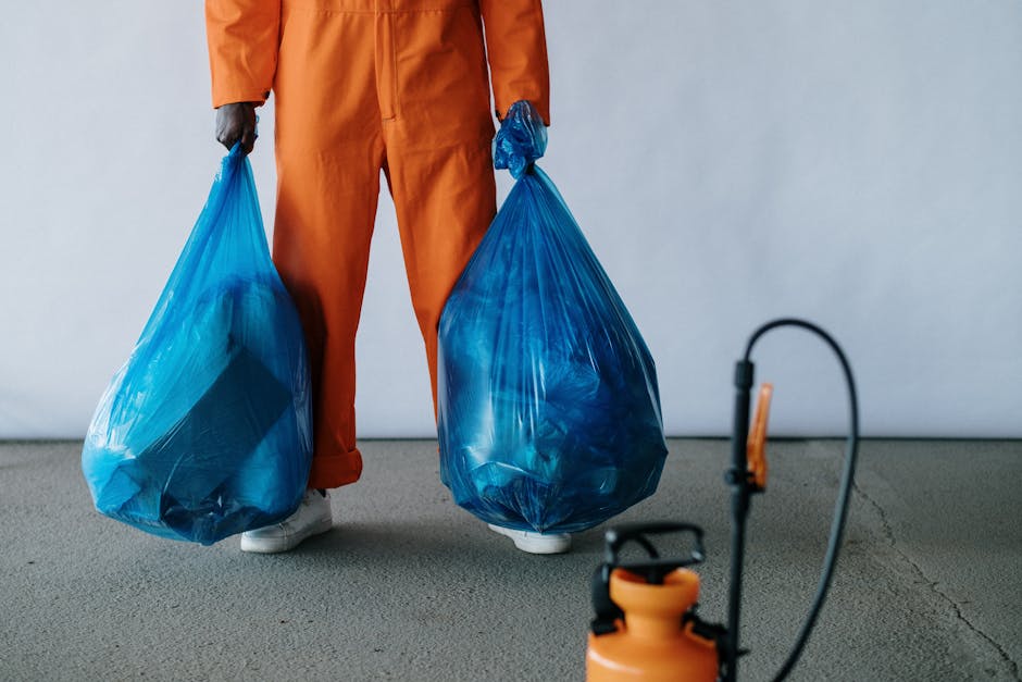 A young man dressed in an orange work jumpsuit stands indoors against a plain light gray background, holding two large blue trash bags filled with waste, with one in each hand. The bags appear to be m