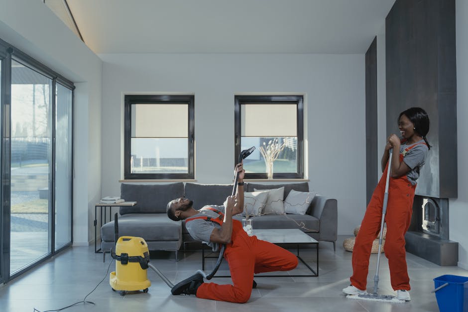 A man and woman dressed in orange cleaning overalls performing domestic cleaning in a modern living room. The man is kneeling on one knee, holding a handheld vacuum cleaner in one hand and a duster in the other, appearing to be in mid-cleaning. The woman is standing, holding a mop with a blue bucket on the floor beside her. The room has a large sliding glass door on the left, bringing in natural light, with a view of an outdoor garden. The walls are painted white with one accent wall in a dark grey color. A grey sofa with white cushions is positioned against the back wall, with two small windows above it. A glass coffee table and a side table with some objects are also visible, and the floor appears clean and well-maintained, reflecting the comprehensive surface cleaning and sanitisation services provided by Finsbury Park Cleaner.