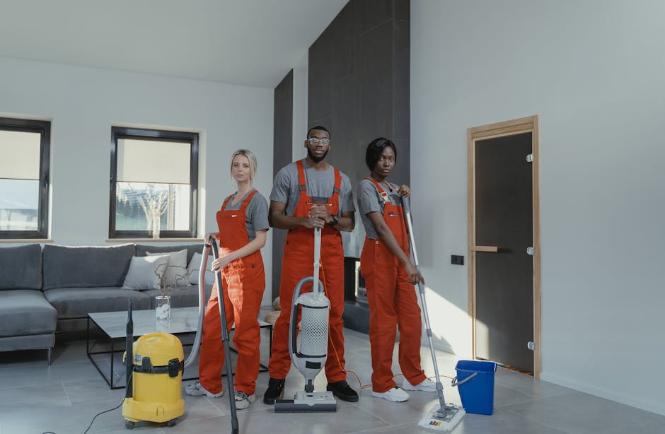 A man and woman dressed in orange cleaning overalls performing domestic cleaning in a modern living room. The man is kneeling on one knee, holding a handheld vacuum cleaner in one hand and a duster in the other, appearing to be in mid-cleaning. The woman is standing, holding a mop with a blue bucket on the floor beside her. The room has a large sliding glass door on the left, bringing in natural light, with a view of an outdoor garden. The walls are painted white with one accent wall in a dark grey color. A grey sofa with white cushions is positioned against the back wall, with two small windows above it. A glass coffee table and a side table with some objects are also visible, and the floor appears clean and well-maintained, reflecting the comprehensive surface cleaning and sanitisation services provided by Finsbury Park Cleaner.