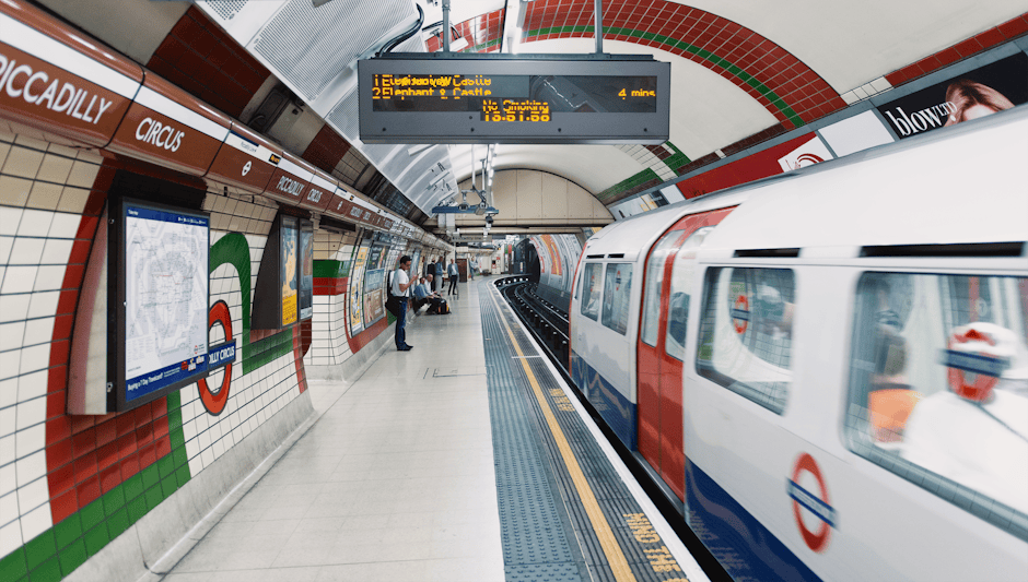 Interior view of Finsbury Park Station subway platform showing clean tiled walls with white, red, and green mosaic patterns, a smooth tiled floor, and a modern train arriving. The platform is illuminated with bright lighting, and a digital schedule display hangs from the curved ceiling, indicating train departure times. Several passengers are waiting, some seated on benches, while others stand near the wall, all maintaining a tidy and hygienic environment. Finsbury Park Cleaner specializes in same-day emergency cleaning and surface sanitisation, ensuring the station remains spotless and well-maintained, as depicted through the pristine condition of the tiled surfaces and spotless floor in this station environment.