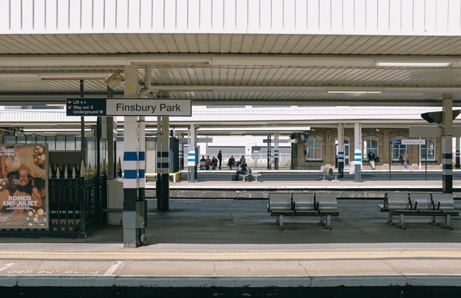 A view of Finsbury Park Station platform with a covered waiting area featuring metal benches and a sign indicating the station name. The platform surface is clean, smooth, and free of litter, with tiled flooring and a yellow safety line along the edge. In the background, passengers are seated on benches or walking along the platform, while station buildings with brick facades and glass windows are visible under the roof structure. The lighting is natural, and the area appears well-maintained, reflecting professional cleaning and upkeep by Finsbury Park Cleaner as part of their deep cleaning and sanitisation services for transport hubs.